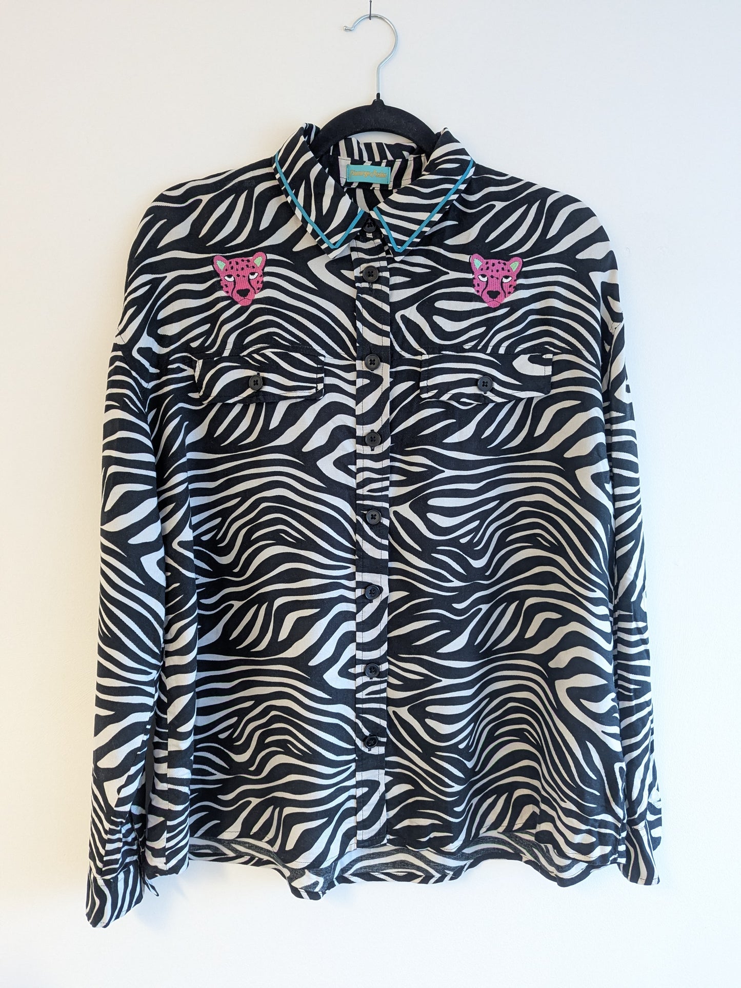 Zebra print shirt with embroidered pink leopard designs on a hanger against a white background. The collar has a contrasting teal ribbon trim around it.