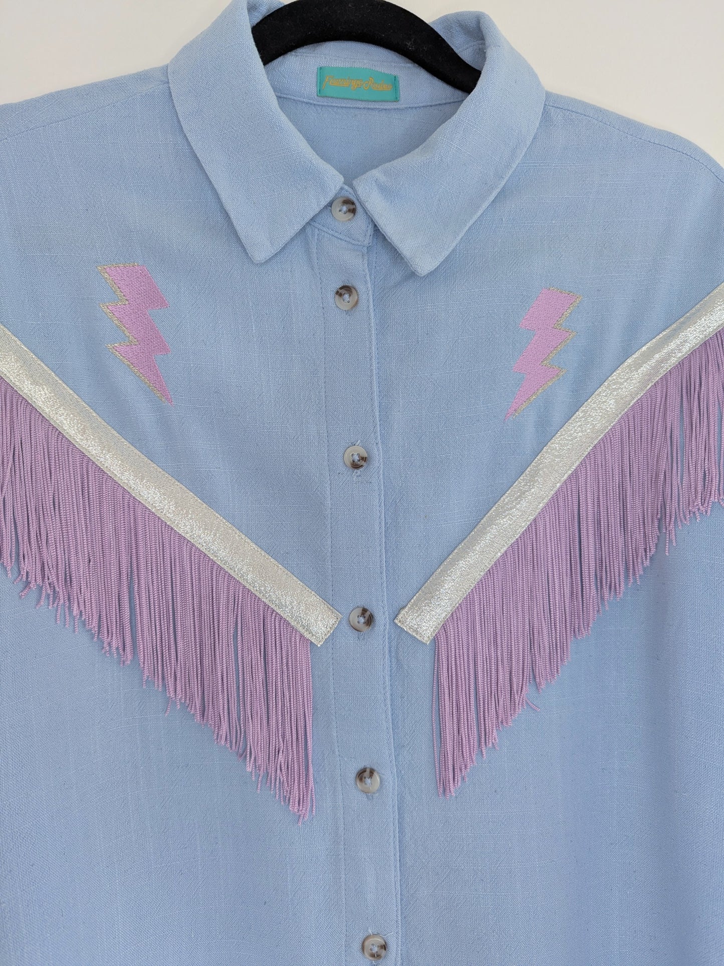 Light blue shirt with lilac lightning bolt embroidered designs and fringe trim on a hanger.