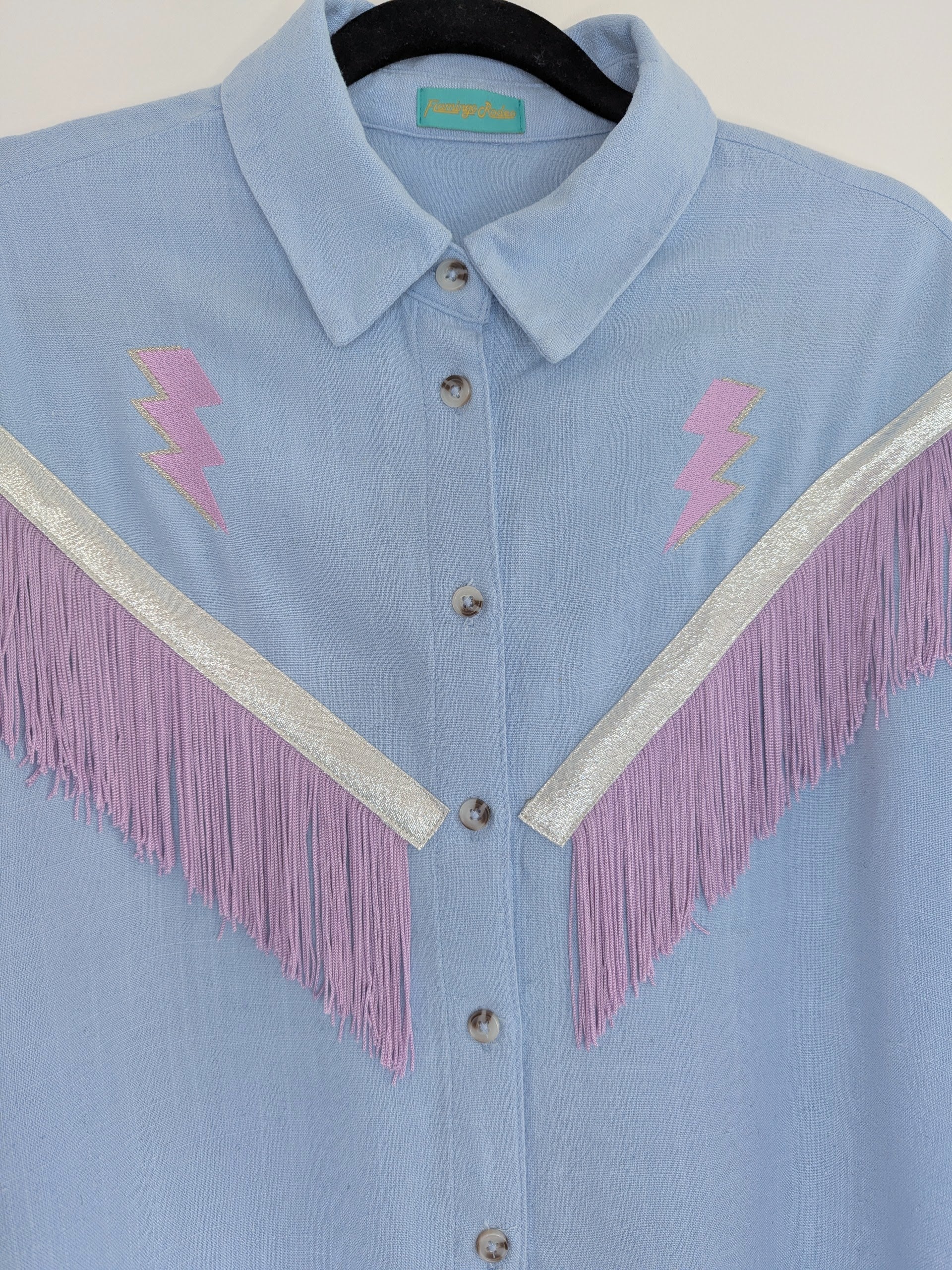 Light blue shirt with lilac lightning bolt embroidered designs and fringe trim on a hanger.