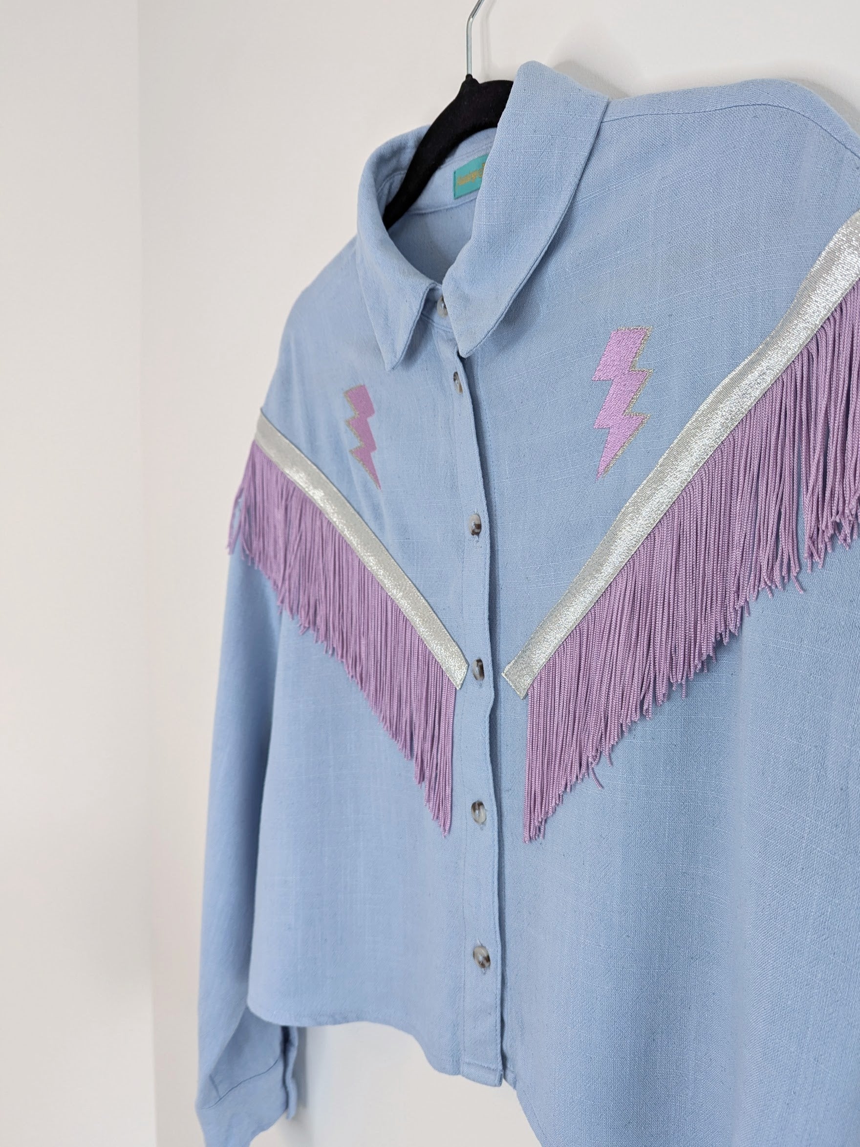 Light blue shirt with lilac and silver fringe details on a hanger against a white background