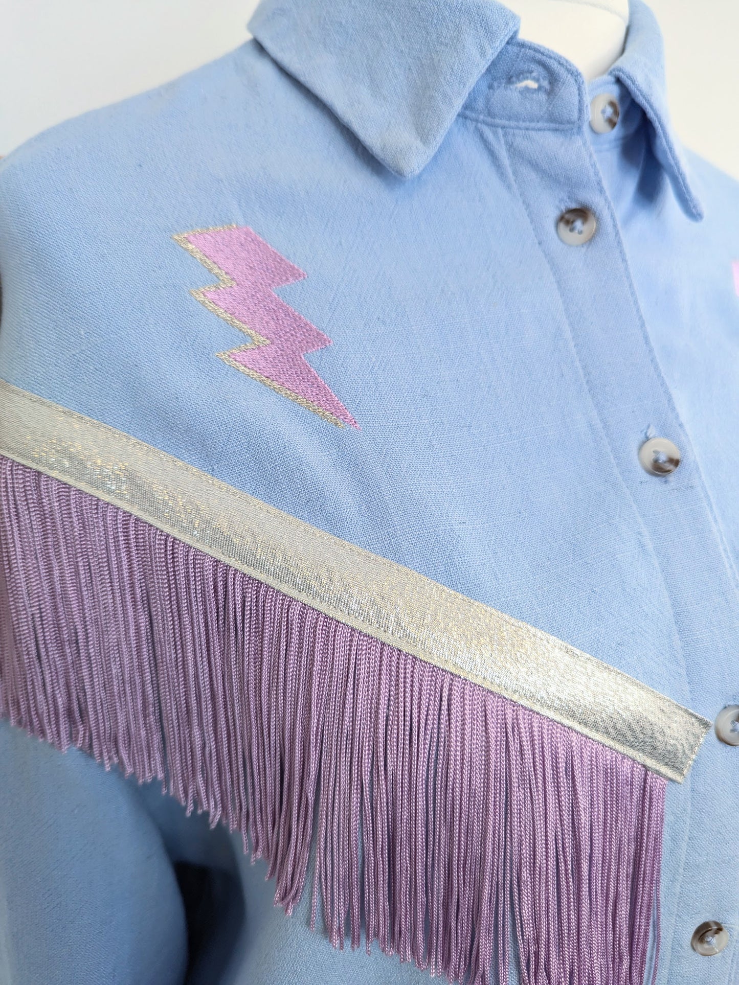 Light blue shirt with lilac and silver embroidered lightning bolts with western style fringe trim on a mannequin against a white background