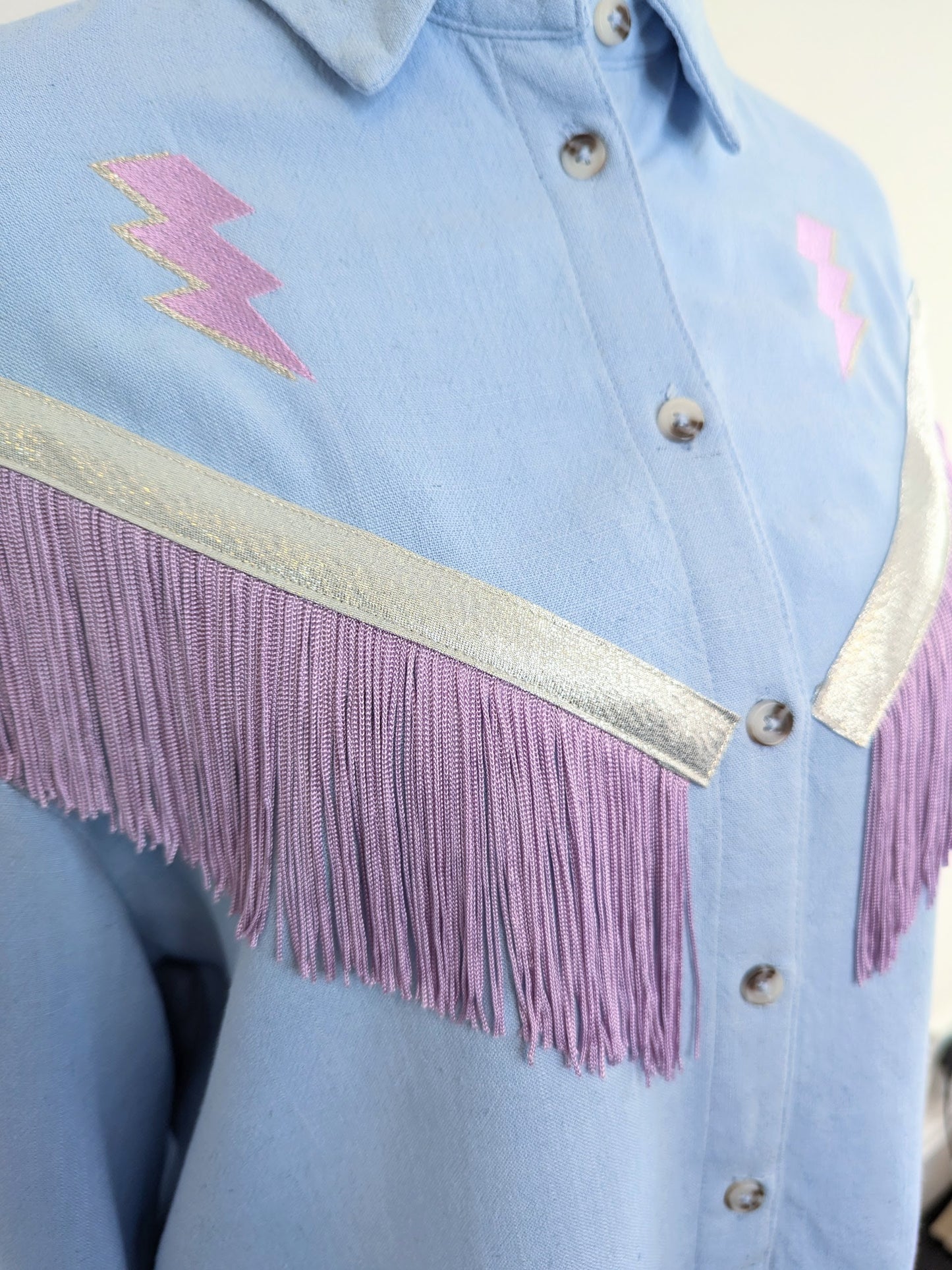 Light blue shirt with lilac and silver embroidered lightning bolts with western style fringe trim on a mannequin against a white background