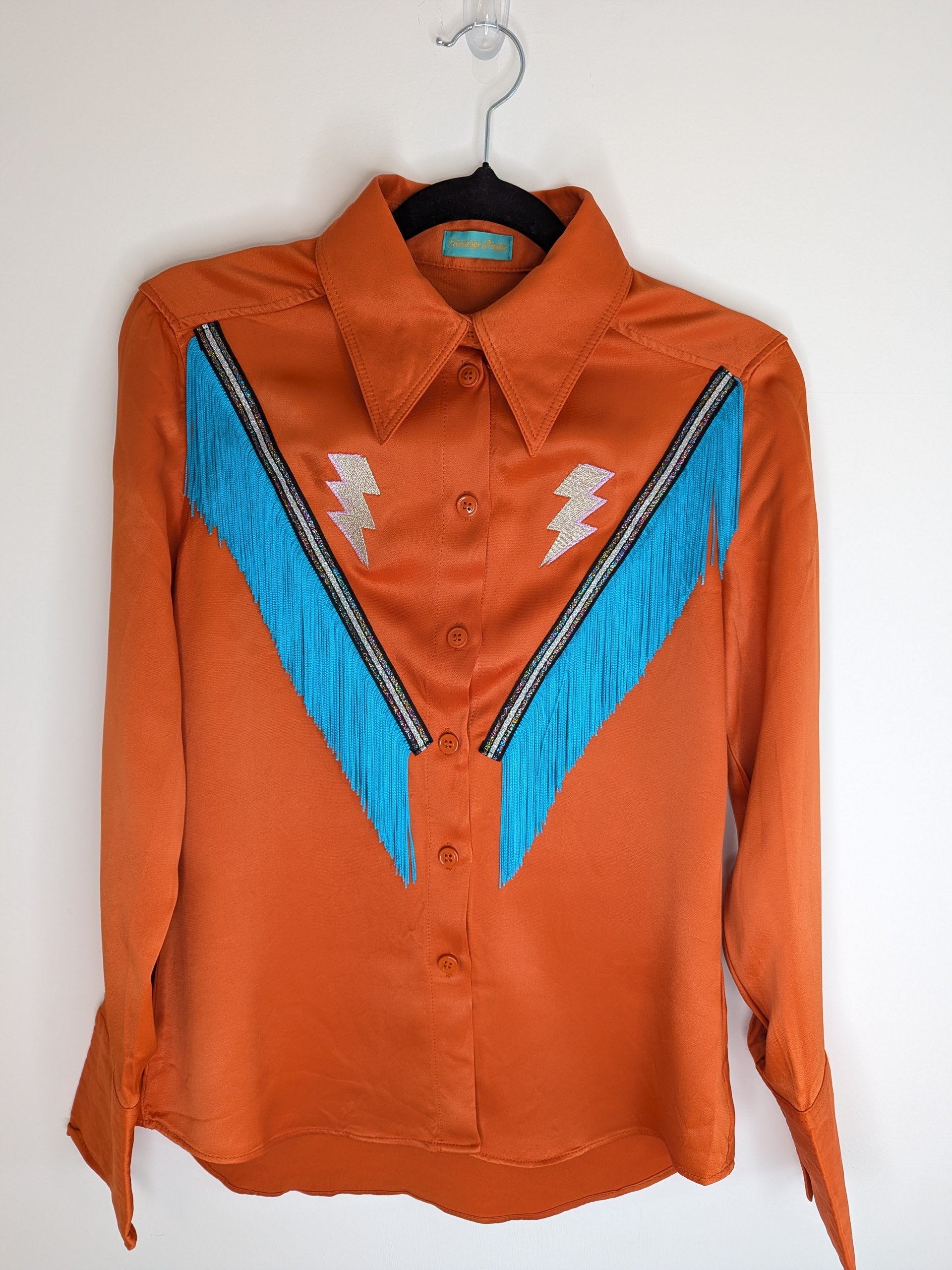 Orange 70's style collar satin shirt with silver lightning bolt embroidery and turquoise fringe trim on a black hanger against a white background