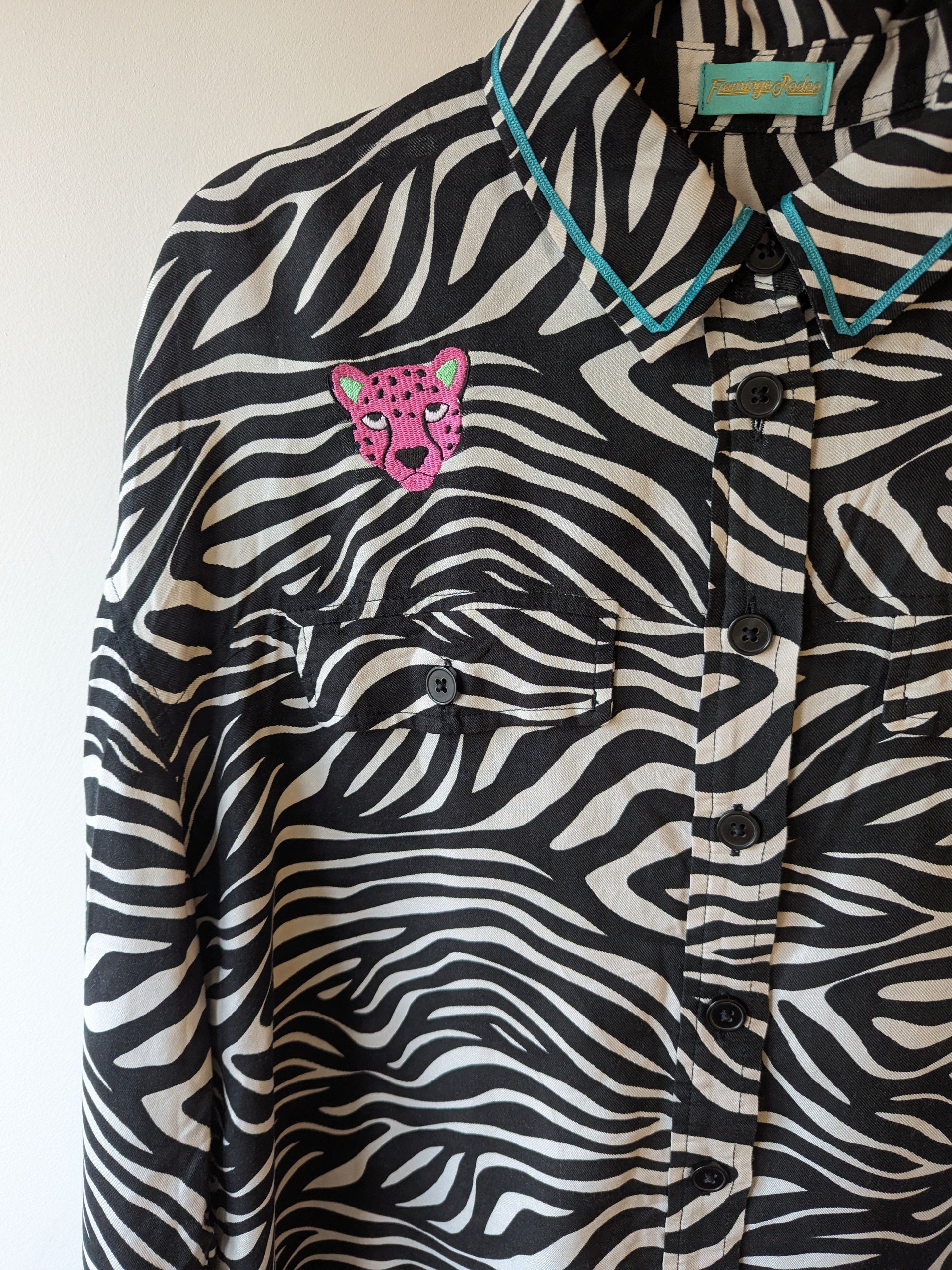Zebra print shirt with embroidered pink leopard designs on a hanger against a white background. The collar has a contrasting teal ribbon trim around it. 