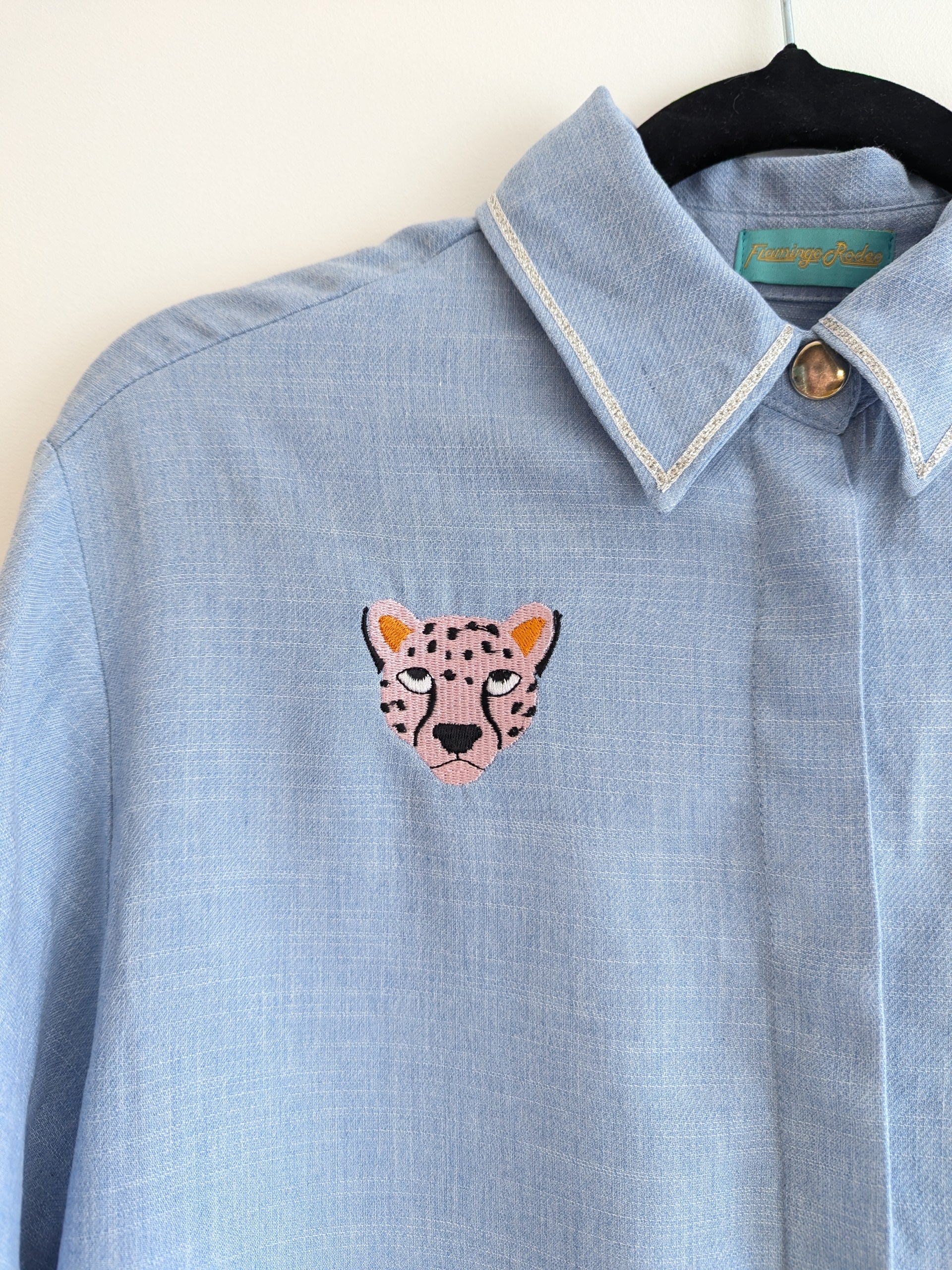 Light blue shirt with contrast silver ribbon trim on the collar and embroidered cheetah faces on a beige background