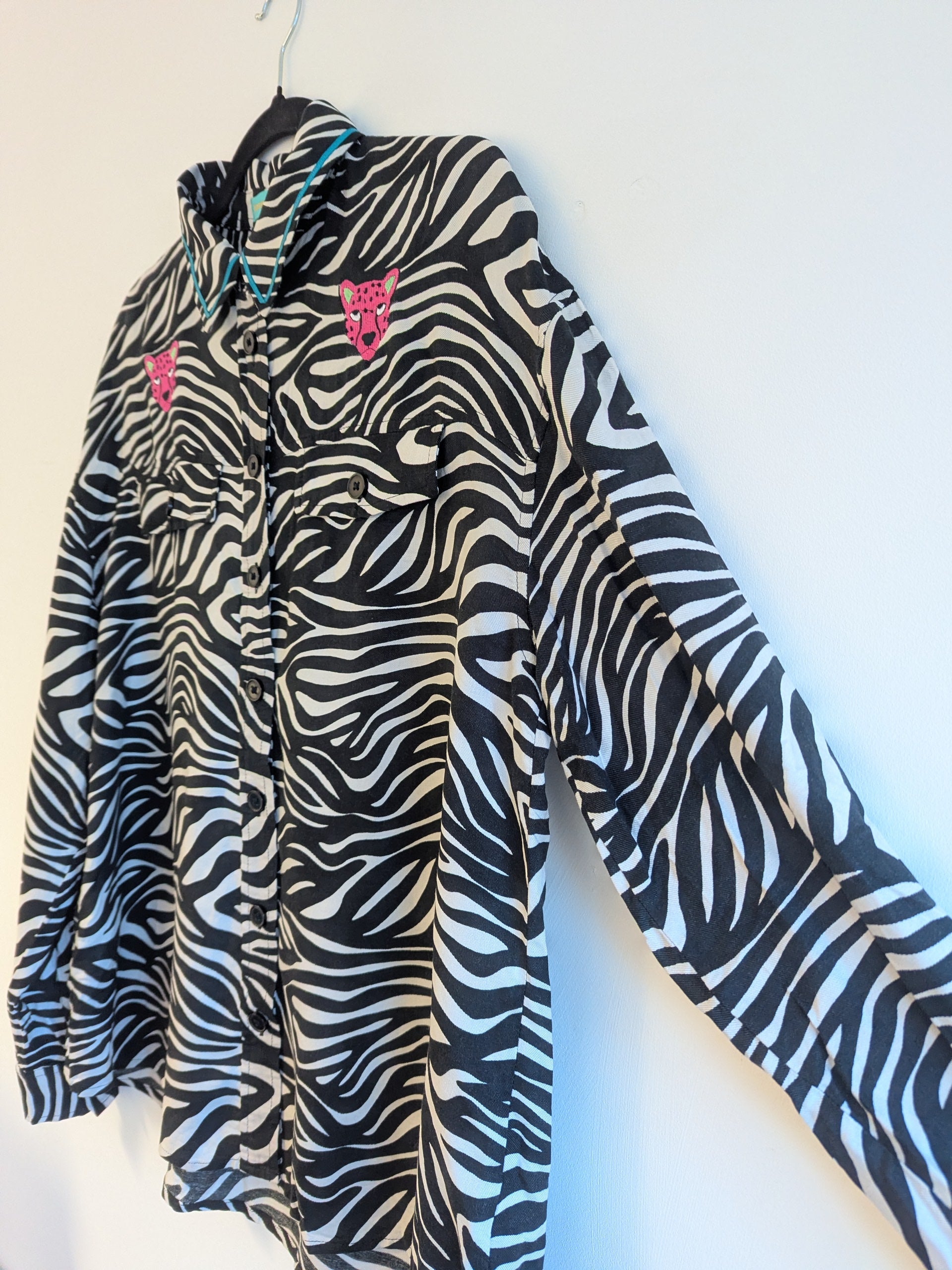 Zebra print shirt with embroidered pink leopard designs on a hanger against a white background. The collar has a contrasting teal ribbon trim around it. 