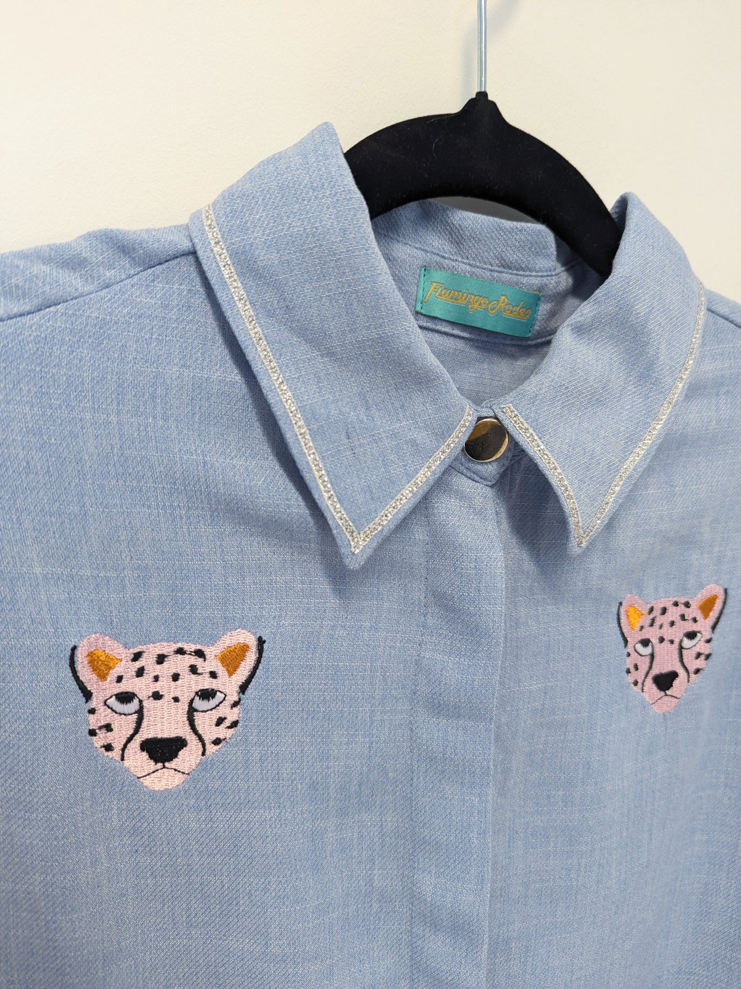 Light blue shirt with contrast silver ribbon trim on the collar and embroidered cheetah faces on a beige background