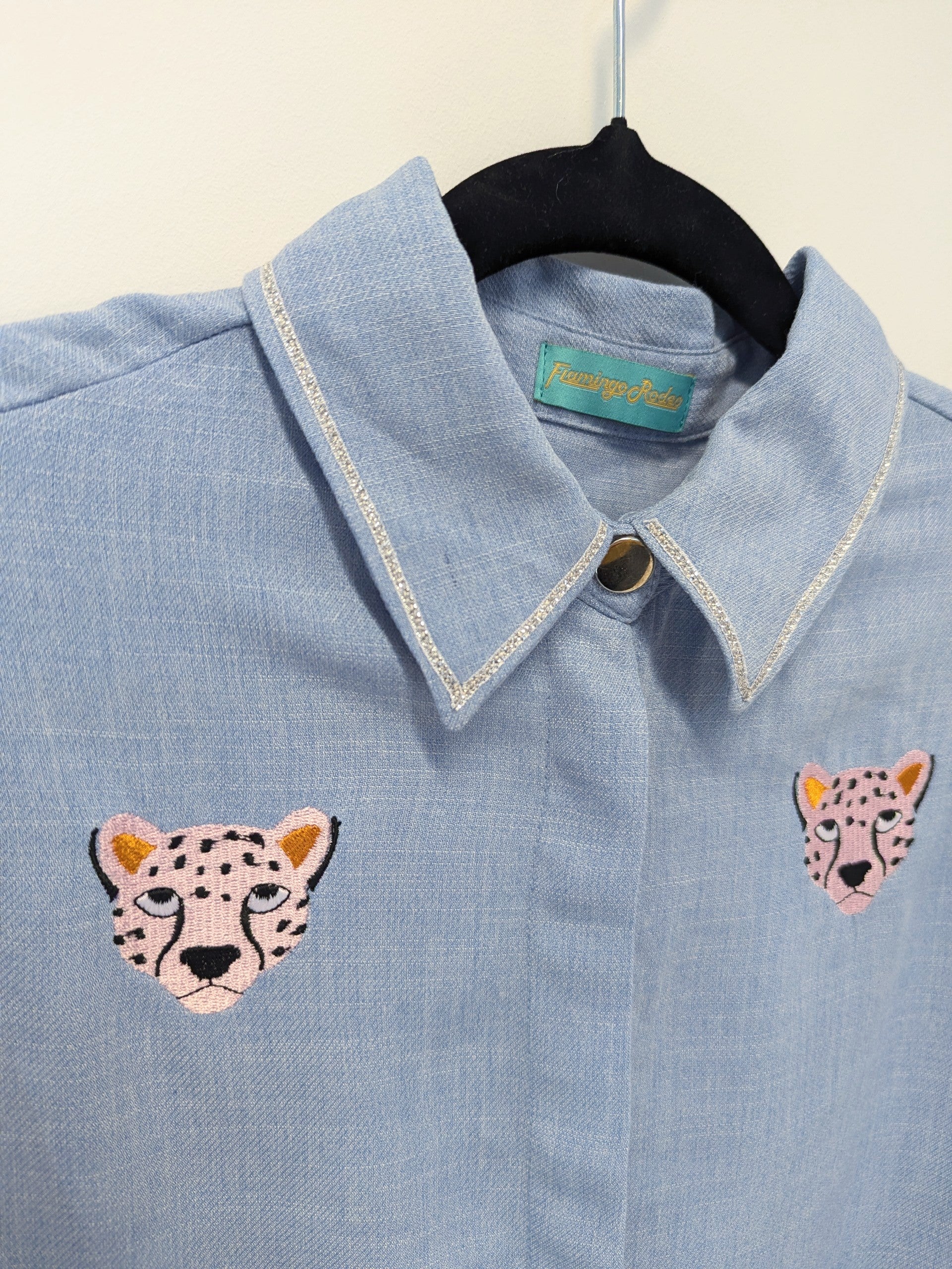 Light blue shirt with contrast silver ribbon trim on the collar and embroidered cheetah faces on a beige background