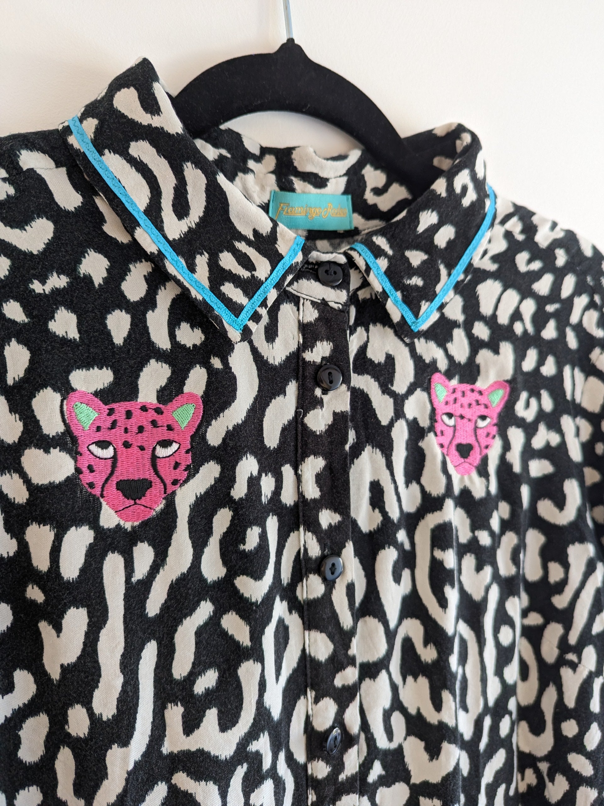Long sleeve black and white animal print shirt with embroidery pink leopard faces on a white background. The shirt has a contrast teal ribbon on the collar. 