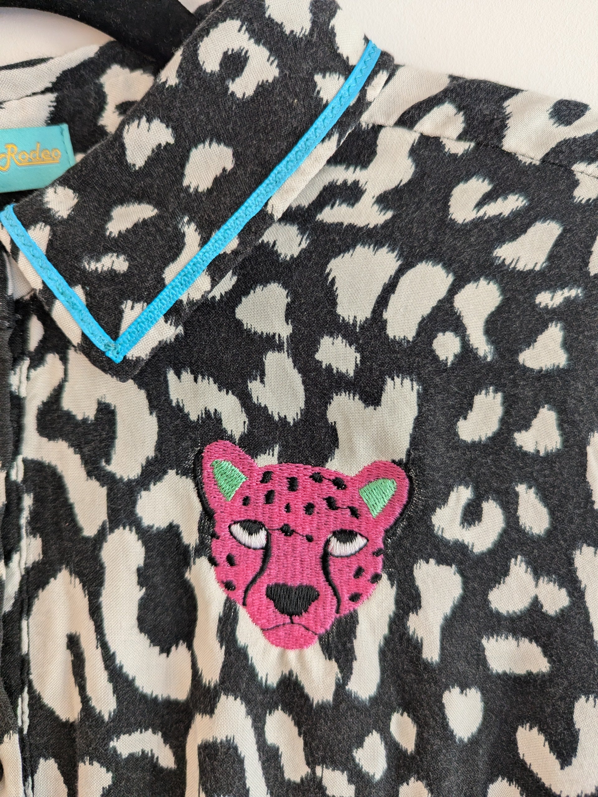 Black and white animal print shirt with embroidery pink leopard faces on a white background. The shirt has a contrast teal ribbon on the collar. 
