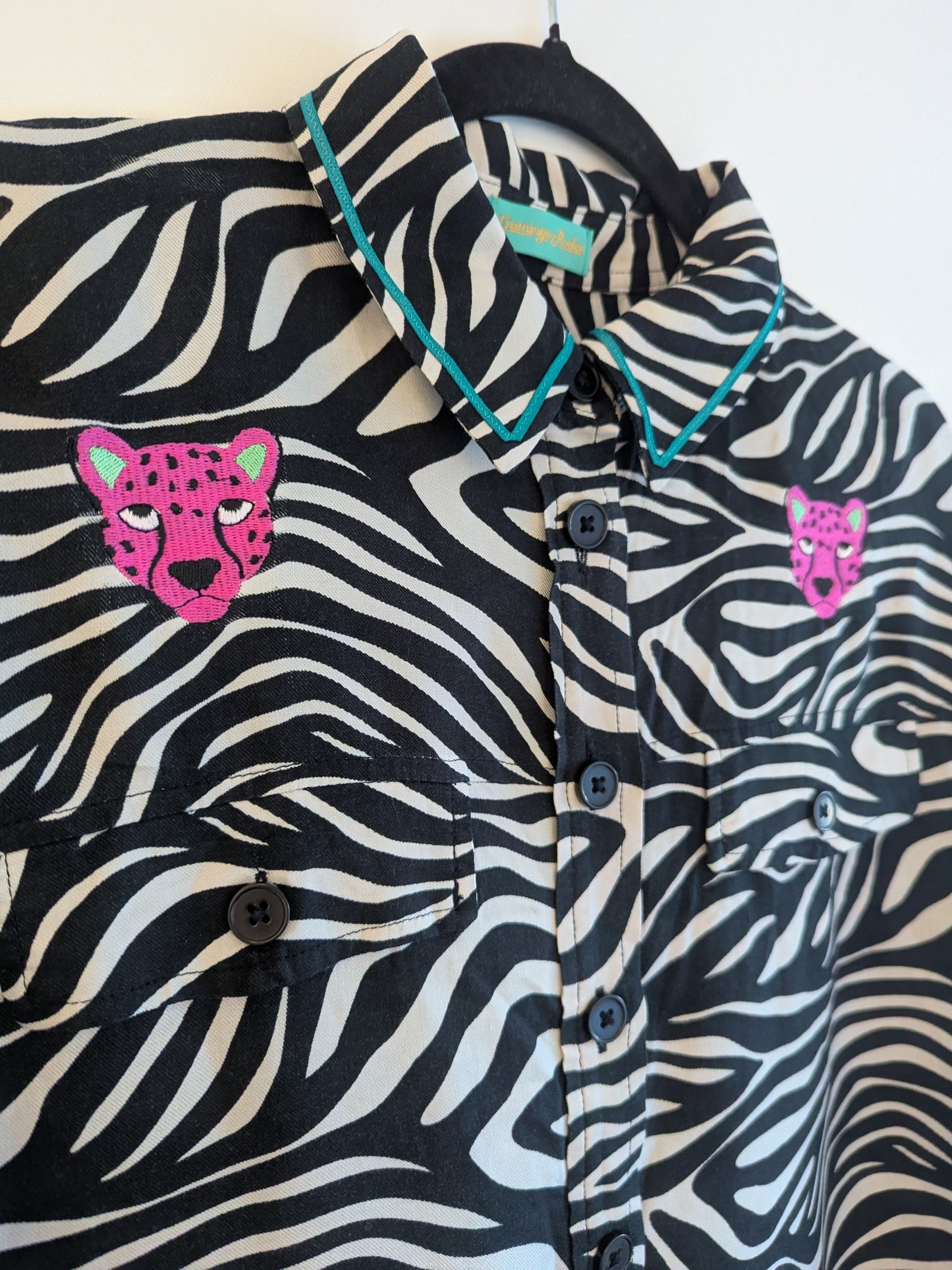 Zebra print shirt with embroidered pink leopard designs on a hanger against a white background. The collar has a contrasting teal ribbon trim around it. 