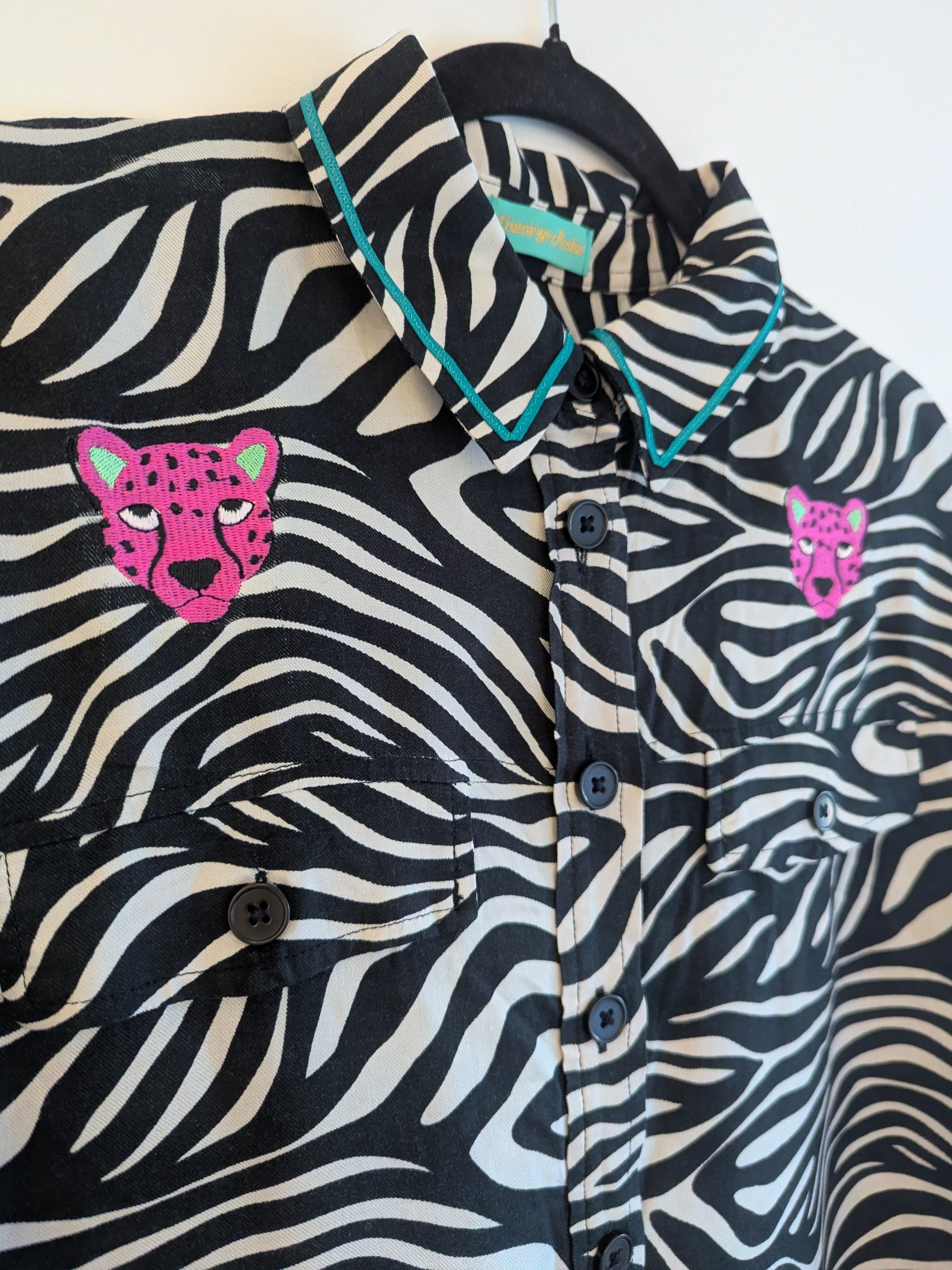 Zebra print shirt with embroidered pink leopard designs on a hanger against a white background. The collar has a contrasting teal ribbon trim around it. 