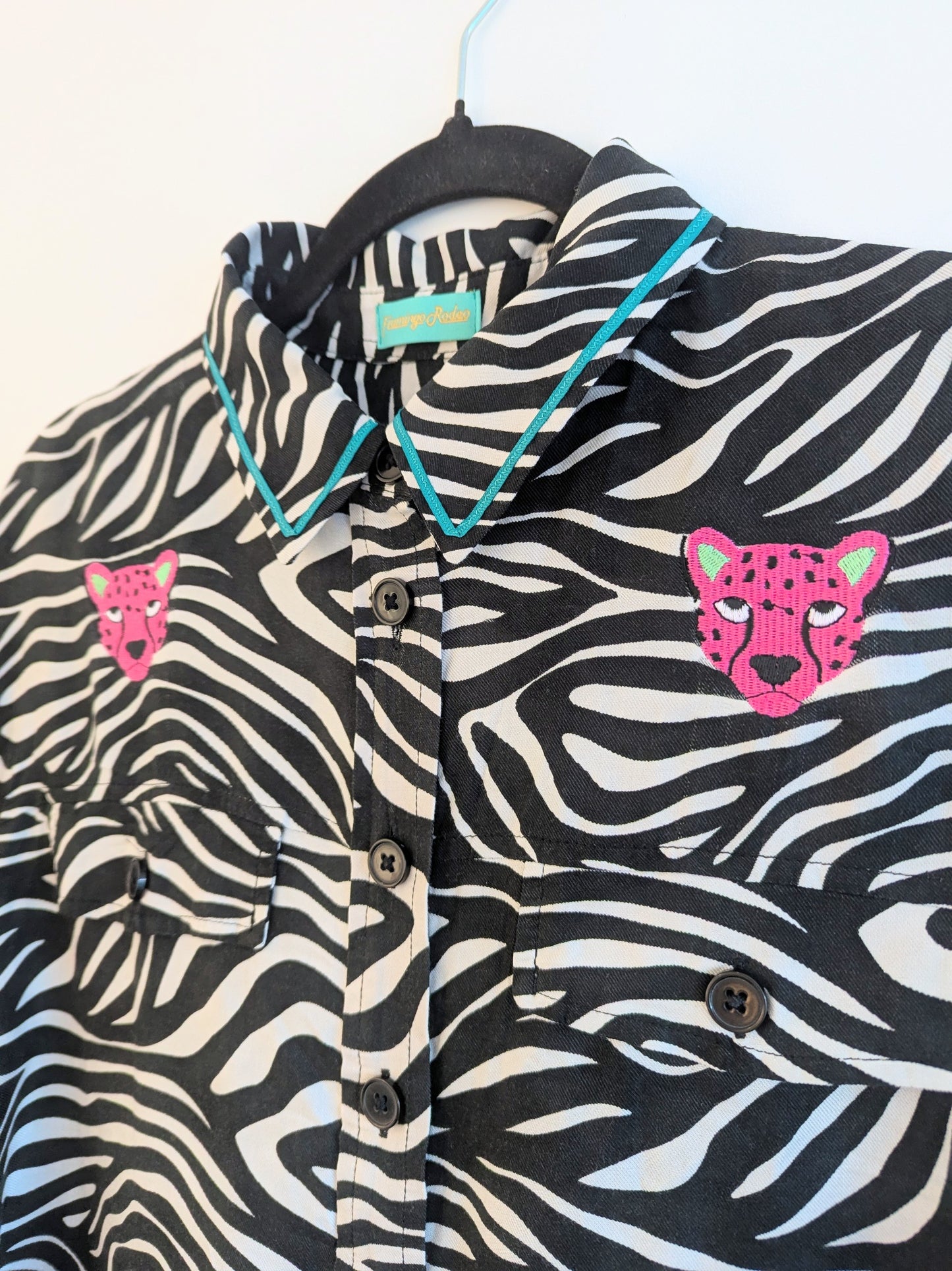 Zebra print shirt with embroidered pink leopard designs on a hanger against a white background. The collar has a contrasting teal ribbon trim around it. 