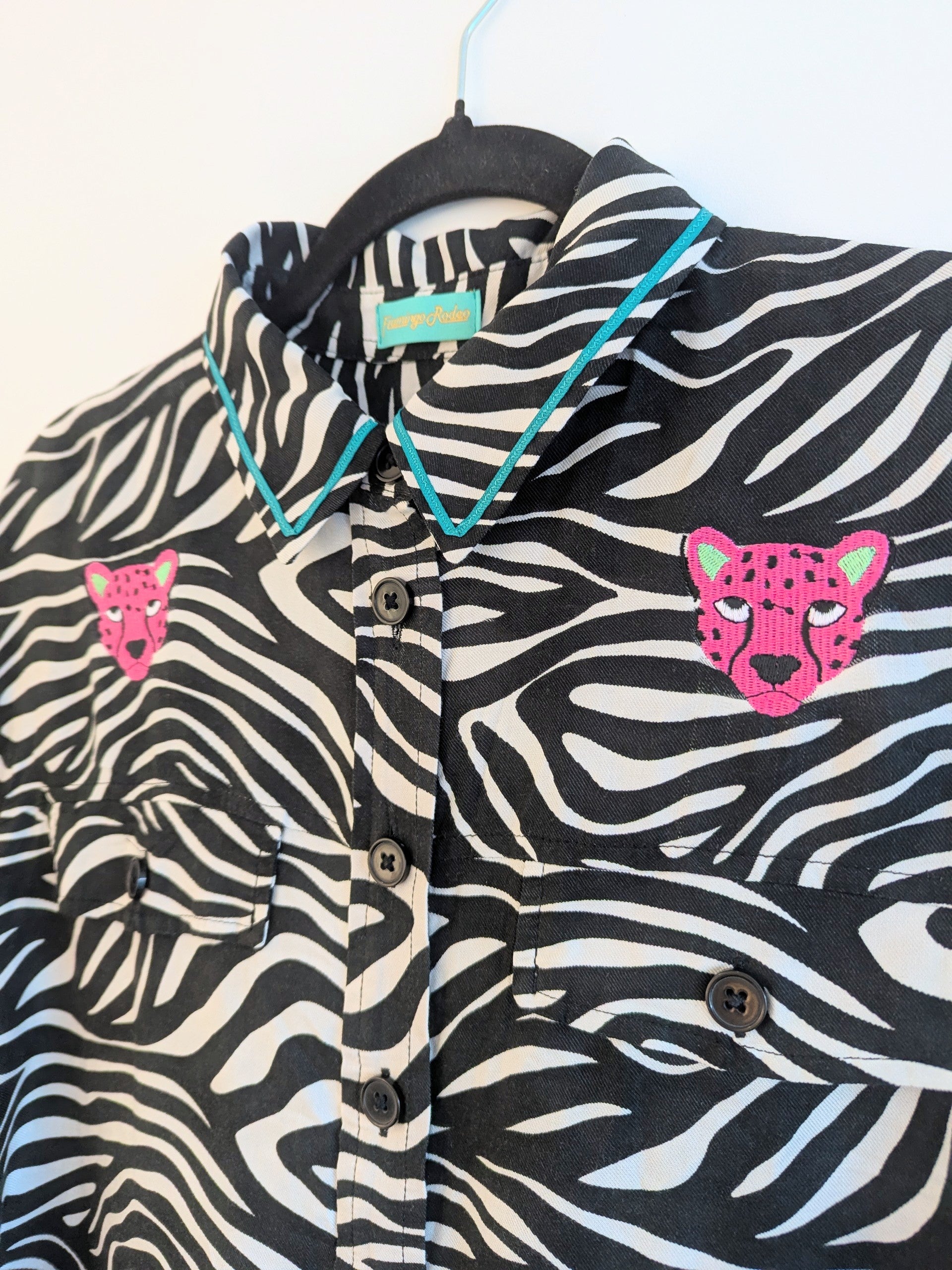 Zebra print shirt with embroidered pink leopard designs on a hanger against a white background. The collar has a contrasting teal ribbon trim around it. 