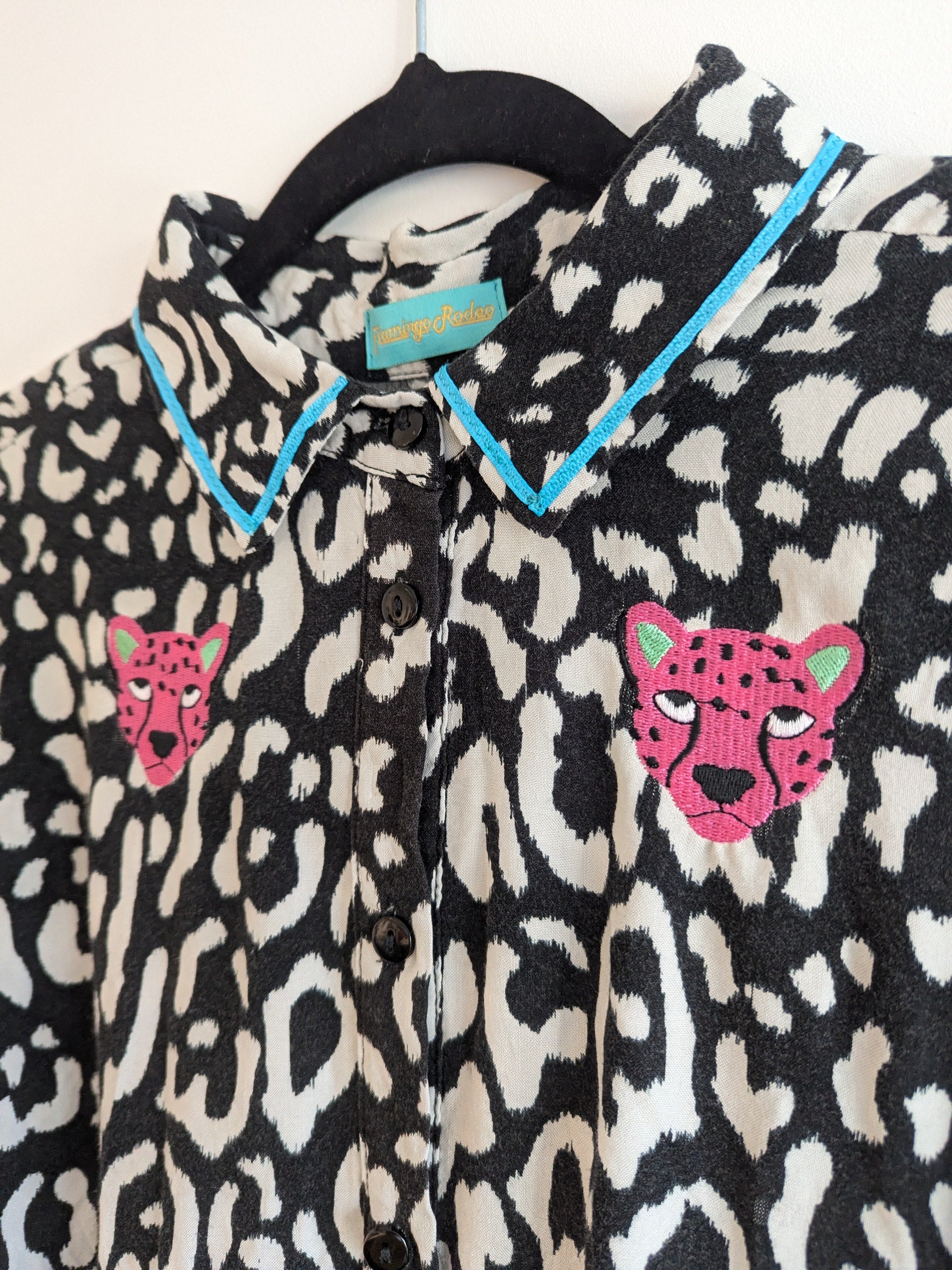 Black and white animal print shirt with embroidery pink leopard faces on a white background. The shirt has a contrast teal ribbon on the collar. 