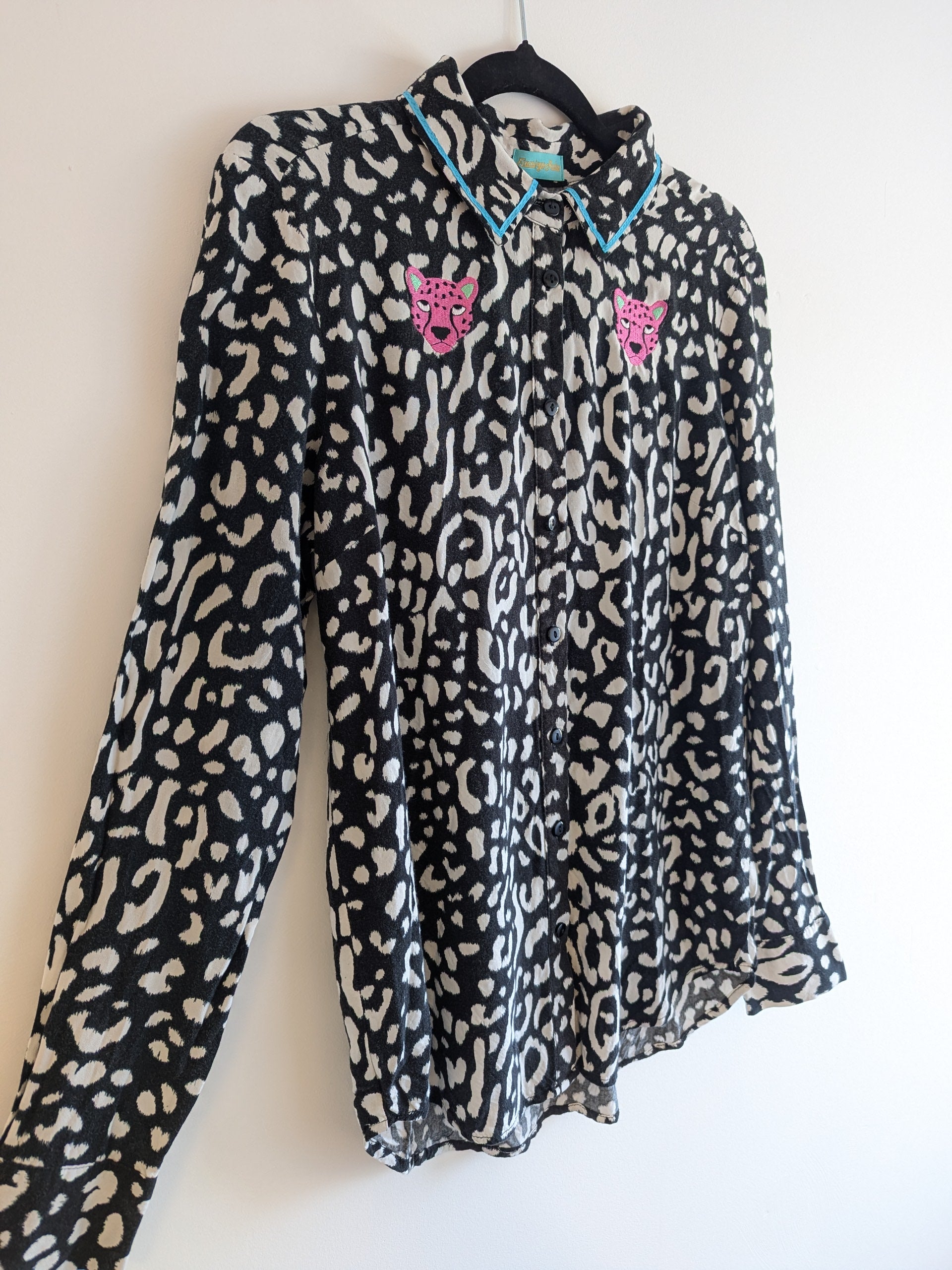 Long sleeve black and white animal print shirt with embroidery pink leopard faces on a white background. The shirt has a contrast teal ribbon on the collar. 