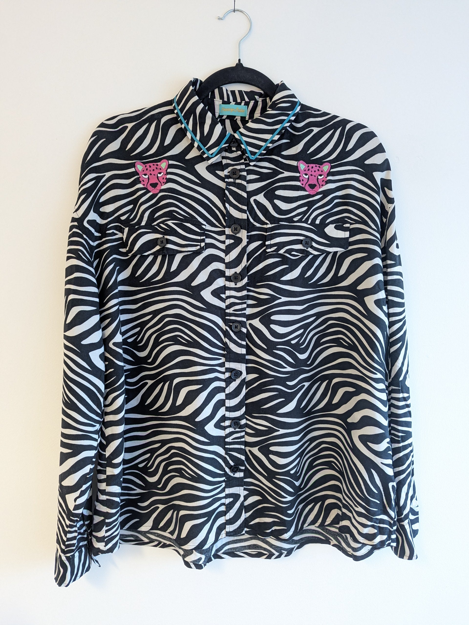 Zebra print shirt with embroidered pink leopard designs on a hanger against a white background. The collar has a contrasting teal ribbon trim around it. 