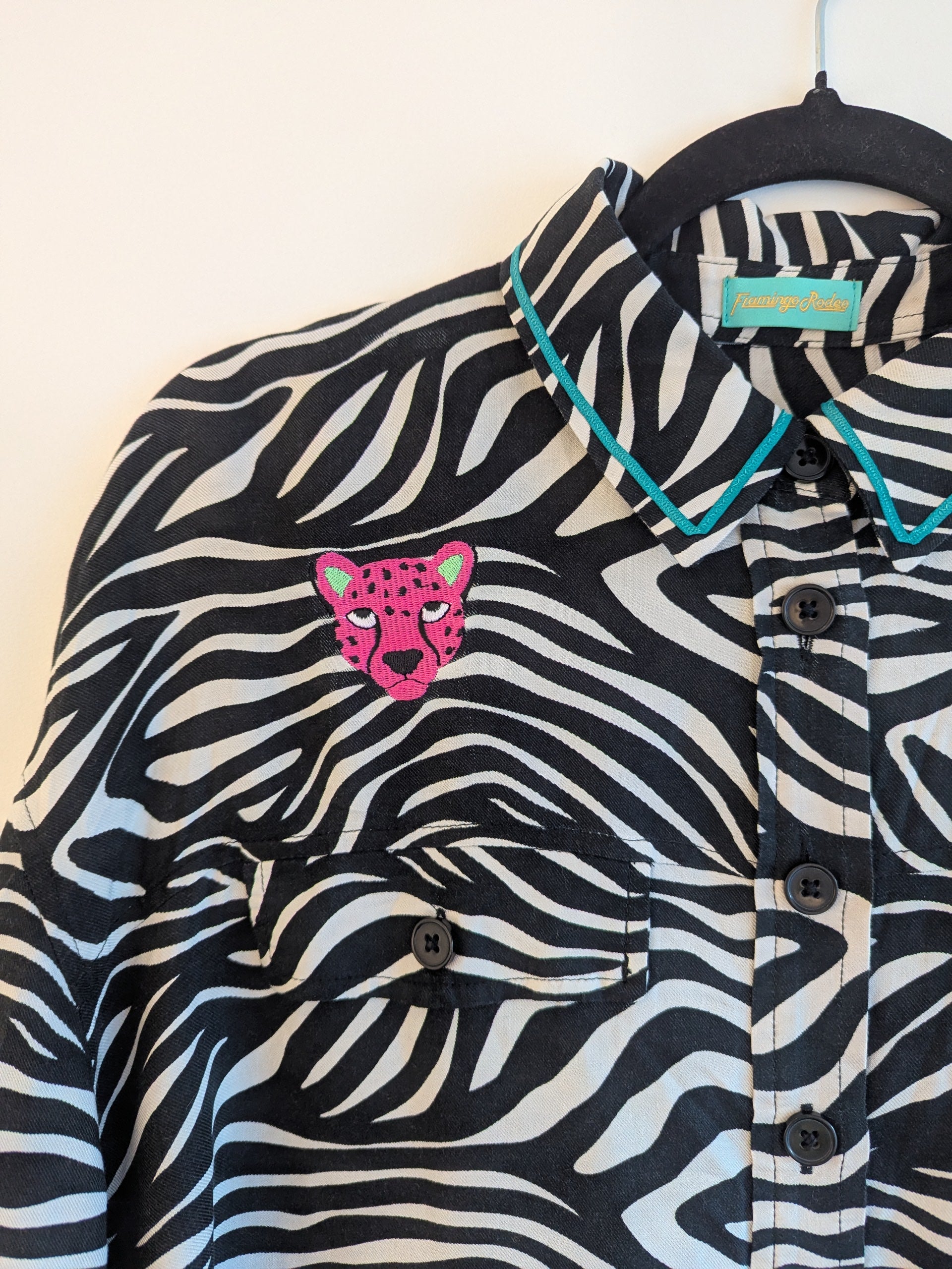 Zebra print shirt with embroidered pink leopard designs on a hanger against a white background. The collar has a contrasting teal ribbon trim around it. 