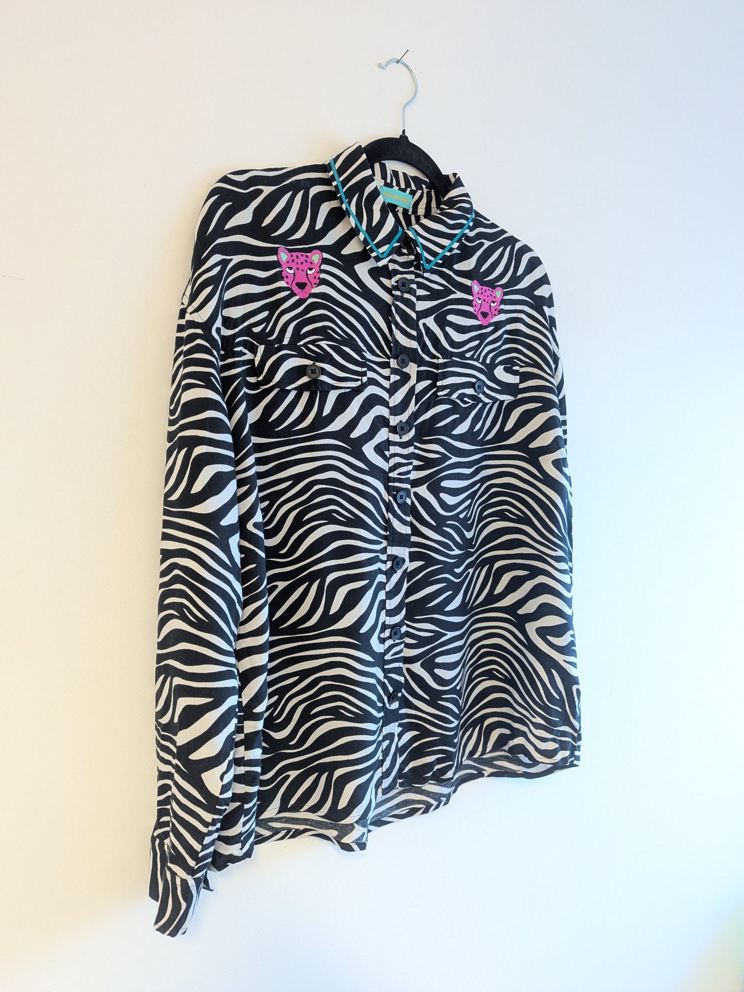 Zebra print shirt with embroidered pink leopard designs on a hanger against a white background. The collar has a contrasting teal ribbon trim around it. 