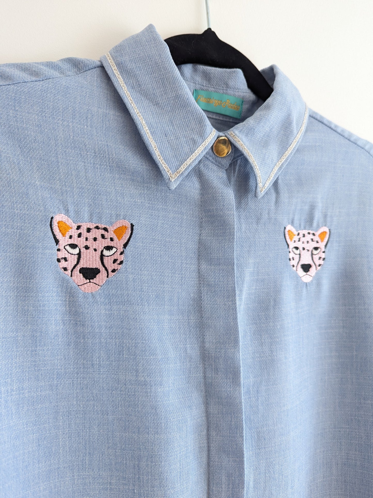 Light blue shirt with contrast silver ribbon trim on the collar and embroidered cheetah faces on a beige background