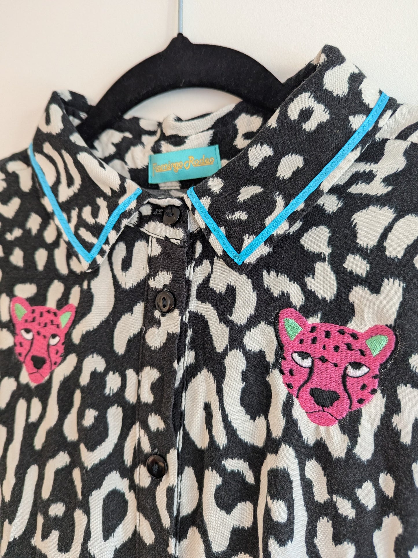 Black and white animal print shirt with embroidery pink leopard faces on a white background. The shirt has a contrast teal ribbon on the collar. 