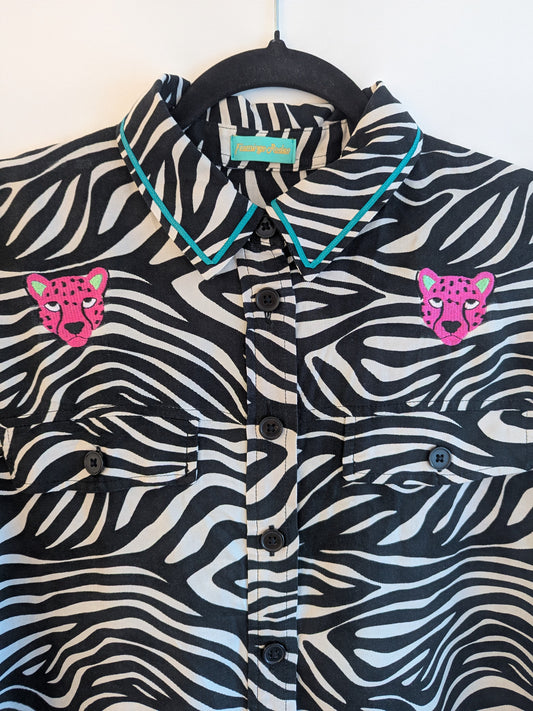 Zebra print shirt with embroidered pink leopard designs on a hanger against a white background. The collar has a contrasting teal ribbon trim around it. 